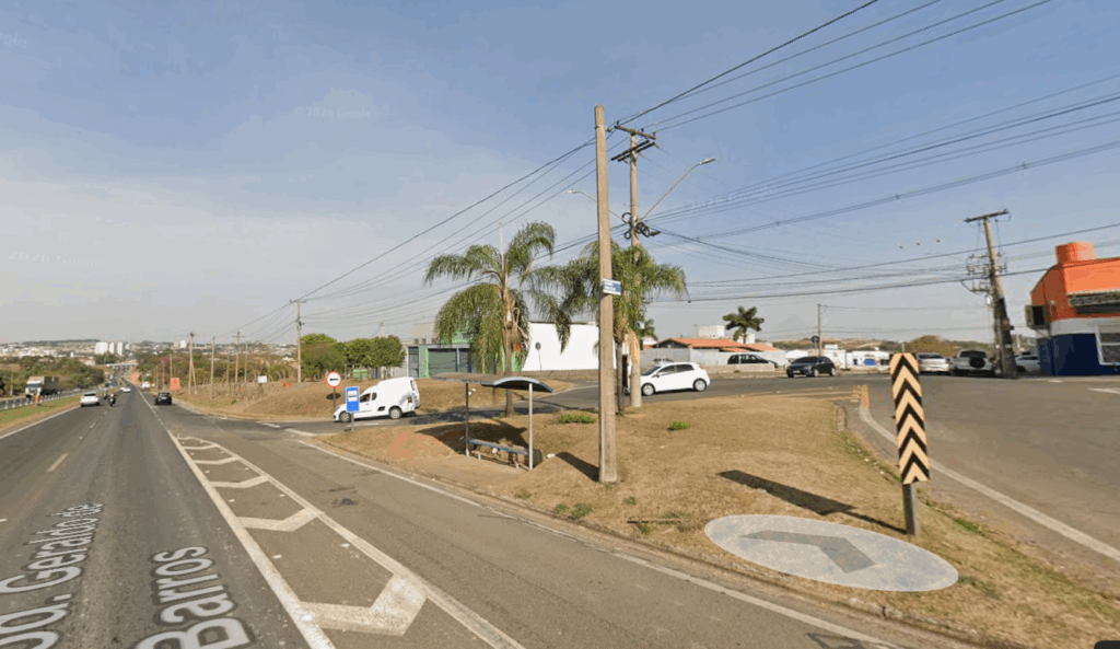 A imagem mostra um cruzamento de ruas em uma área urbana, provavelmente em uma cidade brasileira, dado o estilo das placas e da sinalização. A via principal, chamada "Geraldo de Barros", é larga, com várias faixas e marcações no asfalto, incluindo setas brancas e áreas de sinalização em ziguezague. Há um ponto de ônibus simples, com um abrigo azul e banco, localizado em uma pequena faixa de terra com grama seca, entre as ruas. Próximo ao ponto, há algumas palmeiras e um poste de energia com muitos fios. Um carro branco está fazendo uma conversão à esquerda, enquanto outros veículos estão estacionados ou transitando nas vias. O céu está claro, indicando um dia ensolarado, e as sombras longas sugerem que a foto foi tirada pela manhã ou no final da tarde. A cena transmite uma sensação de um local tranquilo e típico de uma região urbana com infraestrutura básica.