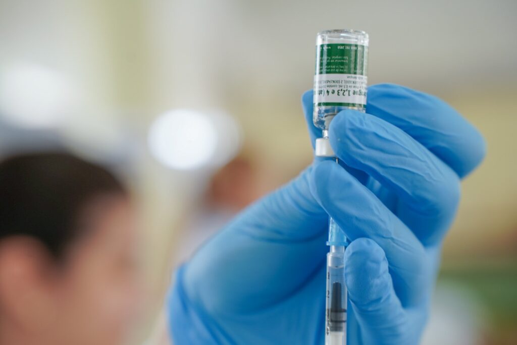 A imagem mostra uma mão vestindo uma luva azul de procedimento segurando um pequeno frasco de vidro de vacina contra a dengue e uma seringa. O frasco está posicionado de cabeça para baixo, com a seringa inserida na tampa de borracha para retirar o líquido. O rótulo do frasco está em português e indica que é uma vacina atenuada contra os quatro sorotipos da dengue (1, 2, 3 e 4). Também há instruções de uso, armazenamento e descarte, como "Vacina de uso único", "Armazenar em geladeira", "Não congelar" e um número de SAC para contato. O fundo está desfocado, sugerindo um ambiente clínico ou hospitalar. A imagem transmite a ideia de um procedimento de vacinação em um contexto de saúde pública.