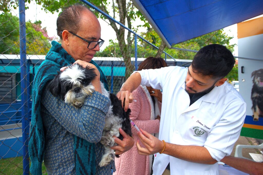 A imagem mostra uma cena veterinária ao ar livre, onde um homem mais velho está segurando com cuidado um cachorro da raça Shih Tzu, que é preto e branco. O homem usa uma camisa azul com estampas e um xale listrado em tons de azul e preto. Ao lado dele, um veterinário jovem, com barba e jaleco branco, está aplicando uma injeção no cachorro. No jaleco do veterinário está escrito "Gustavo Nunes" e "Medicina Veterinária UNESP", indicando sua profissão e instituição. Ao fundo, uma mulher com casaco rosa observa a cena, e há uma estrutura com toldo azul e uma cerca de arame. A luz natural destaca o momento, que parece ser uma vacinação ou tratamento de rotina para o animal. A imagem transmite cuidado, profissionalismo e atenção ao bem-estar do pet.