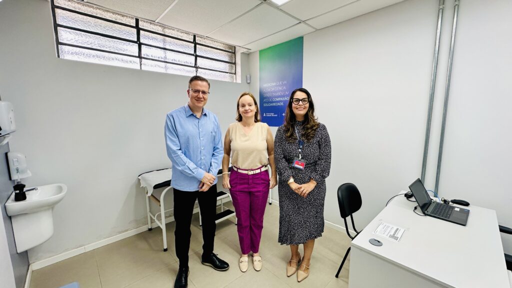 A imagem mostra três pessoas posando em um ambiente que parece ser um consultório médico ou uma sala acadêmica. À esquerda, um homem usa óculos, camisa azul clara e calça preta. No centro, uma mulher loira veste uma blusa sem mangas bege e calças rosa vibrante. À direita, outra mulher com cabelo escuro e ondulado usa óculos grandes, um vestido preto e branco com estampa geométrica e um crachá pendurado no pescoço. Ao fundo, há um cartaz com um gradiente azul para verde contendo a frase em português: "MEDICINA QUE VAI ALÉM DA CIÊNCIA, SENDO TAMBÉM UM ATO DE COMPAIXÃO E SOLIDARIEDADE." Abaixo, está escrito "Universidade Anhembi Morumbi". O ambiente é claro, com paredes brancas, uma pia à esquerda e uma mesa com um laptop à direita. A iluminação é uniforme e o espaço parece limpo e organizado.