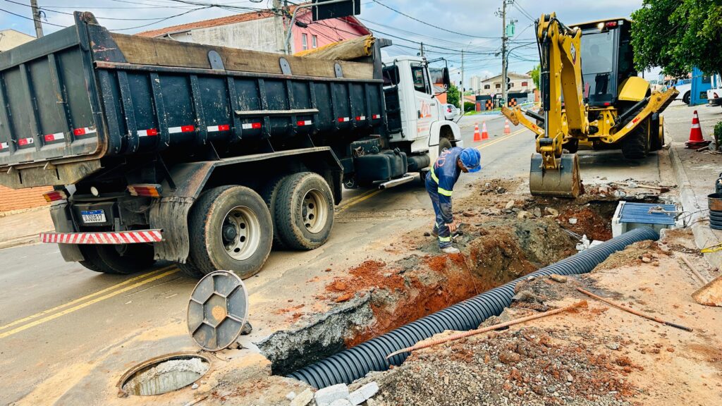 A imagem mostra uma cena de construção em uma estrada durante o dia. No centro, há uma vala profunda aberta no asfalto, onde está sendo instalada uma grande tubulação preta corrugada, provavelmente para drenagem. Um trabalhador, vestido com uniforme azul e capacete, está agachado próximo à vala, aparentemente realizando algum trabalho manual. À esquerda da imagem, há um caminhão caçamba preto com cabine branca, estacionado próximo à vala, com suas rodas traseiras quase encostando na borda da escavação. À direita, um retroescavadeira amarela da marca CAT está posicionada ao lado da vala, com a caçamba apoiada no chão perto de uma grade de drenagem. O chão ao redor da vala é uma mistura de asfalto, terra vermelha e cascalho. Ao fundo, é possível ver casas residenciais, postes de energia elétrica e cones de sinalização laranja que delimitam a área da obra. A iluminação é natural, indicando um dia claro. No geral, a imagem transmite o ambiente típico de uma obra de infraestrutura urbana, com máquinas pesadas e trabalho manual integrados para a instalação de tubulações subterrâneas.
