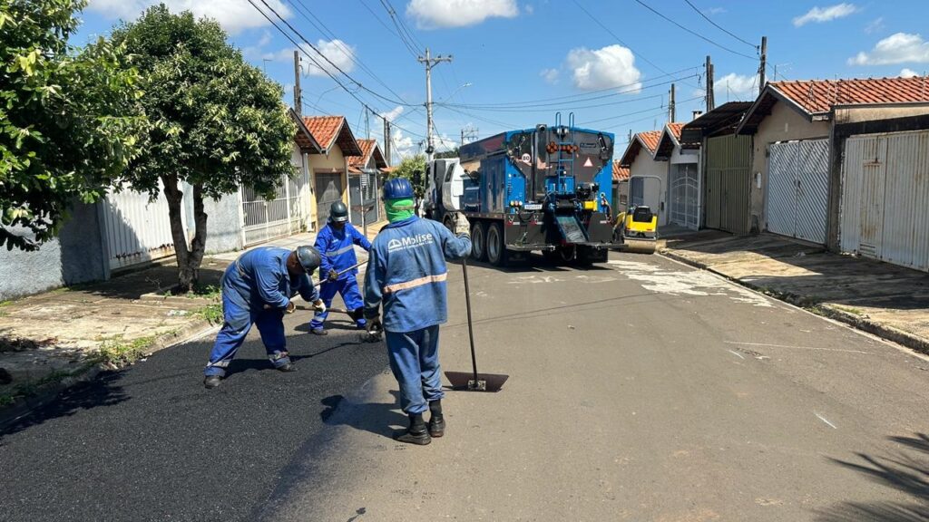 A imagem mostra uma equipe de trabalhadores realizando a pavimentação de uma rua em um bairro residencial. Três homens, vestidos com uniformes azuis e capacetes de segurança, estão concentrados no trabalho: um deles está abaixado usando uma pá, outro está com uma ferramenta na mão, e o terceiro, de costas para a câmera, segura um instrumento longo que parece ser um ancinho para espalhar o asfalto. O asfalto recém-aplicado é escuro e ainda úmido, contrastando com a parte mais antiga e desgastada da via. Ao fundo, há um caminhão grande, azul e branco, provavelmente usado para distribuir o asfalto, e uma pequena máquina compactadora amarela. A rua é estreita e cercada por casas com telhados vermelhos e portões brancos ou cinzas. No céu azul, há algumas nuvens brancas, e postes de energia com muitos fios atravessam a cena. Uma árvore verde está posicionada à esquerda da imagem, trazendo um toque natural ao ambiente urbano. No canto da imagem, aparece a inscrição "Molise Serviços e Construções Ltda", indicando possivelmente a empresa responsável pela obra. A iluminação é forte, sugerindo que a foto foi tirada durante o dia, com sombras bem definidas no chão.