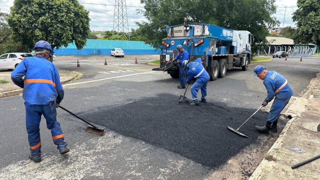A imagem mostra uma equipe de trabalhadores realizando manutenção em uma via urbana. Quatro operários, todos vestidos com uniformes azuis e usando capacetes de segurança, estão aplicando e nivelando uma nova camada de asfalto em um trecho da rua. Um dos trabalhadores varre a área ao redor, enquanto os outros três manipulam ferramentas para espalhar e alisar o material recém-colocado. Ao fundo, há um caminhão azul e branco que parece ser o veículo de apoio para o transporte do asfalto, com inscrições como "Molise Serviços de Construção Ltda." e "Prefeitura de Vitória da Conquista". A palavra "PARE" está pintada no chão, indicando uma faixa de parada próxima. O cenário é ao ar livre, em uma área urbana com algumas árvores e construções ao fundo, sob um céu claro. A cena transmite uma atividade de reparo viário em andamento, mostrando o trabalho manual e coordenado da equipe.