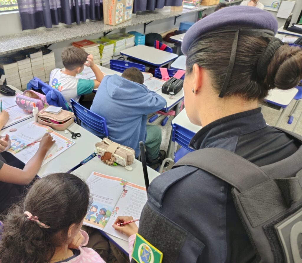 A imagem mostra uma cena dentro de uma sala de aula, onde uma policial feminina, vestindo uniforme azul e um gorro roxo, está de pé, observando atentamente uma aluna que está sentada à sua frente, concentrada em seu material de estudo. A policial tem o cabelo preso em um coque e uma insígnia visível na manga do uniforme, indicando que pertence à polícia militar de algum estado brasileiro. Na sala, várias crianças estão sentadas em carteiras brancas com cadeiras azuis, todas focadas em seus livros abertos. Em destaque, um dos livros parece tratar do programa PROERD (Programa Educacional de Resistência às Drogas), sugerido pelo texto visível nas páginas. Ao fundo, há uma estante com livros e caixas, uma delas com a palavra "DIVERSÕES" escrita. A luz natural entra pela janela, iluminando suavemente o ambiente. A atmosfera transmite um momento de aprendizado e interação entre a polícia e os alunos, possivelmente parte de uma ação educativa na escola.