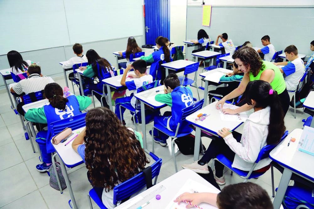 A imagem mostra uma sala de aula cheia de estudantes sentados em carteiras individuais organizadas em fileiras. A maioria dos alunos veste uniformes escolares azuis e brancos. O ambiente é iluminado por luz natural e artificial, com paredes brancas e móveis predominantemente azuis e brancos, o que transmite uma sensação de limpeza e organização. No lado direito da imagem, uma professora de cabelos cacheados, vestindo uma blusa verde-limão sem mangas e calça preta, está inclinada, auxiliando um dos alunos. Os estudantes parecem concentrados em suas atividades, com papéis e materiais escolares sobre as carteiras. A perspectiva da foto é de um ângulo alto, mostrando a sala de trás para frente, o que permite visualizar bem o número de alunos e a disposição do ambiente. No geral, a cena representa um momento típico de estudo e aprendizado em uma escola, com uma interação ativa entre professora e alunos em um ambiente organizado e acolhedor.