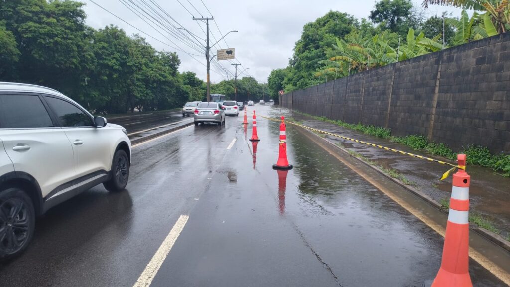A imagem mostra uma rua asfaltada e molhada, provavelmente após uma chuva, com cones de trânsito laranja alinhados ao longo de uma das faixas, indicando uma área alagada. O cenário é urbano ou suburbano, com uma parede de tijolos cinza à direita e árvores verdes à esquerda. O céu está nublado e cinzento, reforçando a sensação de tempo chuvoso. Na lateral da rua, há uma placa amarela com a silhueta de um animal e o texto em português: "ATENÇÃO ANIMAIS NA PISTA REDUZA A VELOCIDADE", alertando os motoristas para a presença de animais na via e pedindo cautela. Vários carros estão na estrada, incluindo um SUV branco em primeiro plano à esquerda. A cena transmite uma sensação de cuidado e precaução devido à combinação do alagamento e da presença possível de animais na pista.