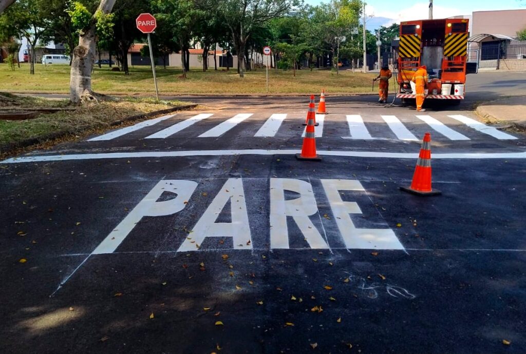 A imagem mostra uma cena de manutenção viária em uma rua, onde está sendo pintada uma faixa de parada para pedestres. No chão, em letras grandes e brancas, está escrita a palavra "PARE", que significa "pare" em português, indicando um ponto de parada para veículos. Ao redor da palavra, há algumas folhas secas espalhadas e marcas de giz, provavelmente usadas para orientação na pintura. À frente da palavra, há uma linha branca e uma faixa de pedestres recém-pintadas, protegidas por três cones de trânsito laranja com faixas refletivas. No fundo da imagem, há um caminhão de manutenção laranja com faixas em zigue-zague amarelas e pretas na parte traseira. Dois trabalhadores vestidos com macacões laranjas de alta visibilidade estão próximos ao caminhão, provavelmente responsáveis pela pintura das sinalizações no chão. A cena ocorre em um dia ensolarado, com árvores verdes e um gramado ao lado da rua, além de uma placa de trânsito vermelha em forma de octógono com a palavra "PARE" escrita, reforçando a sinalização de parada. A imagem transmite um ambiente de trabalho urbano voltado para a segurança no trânsito.