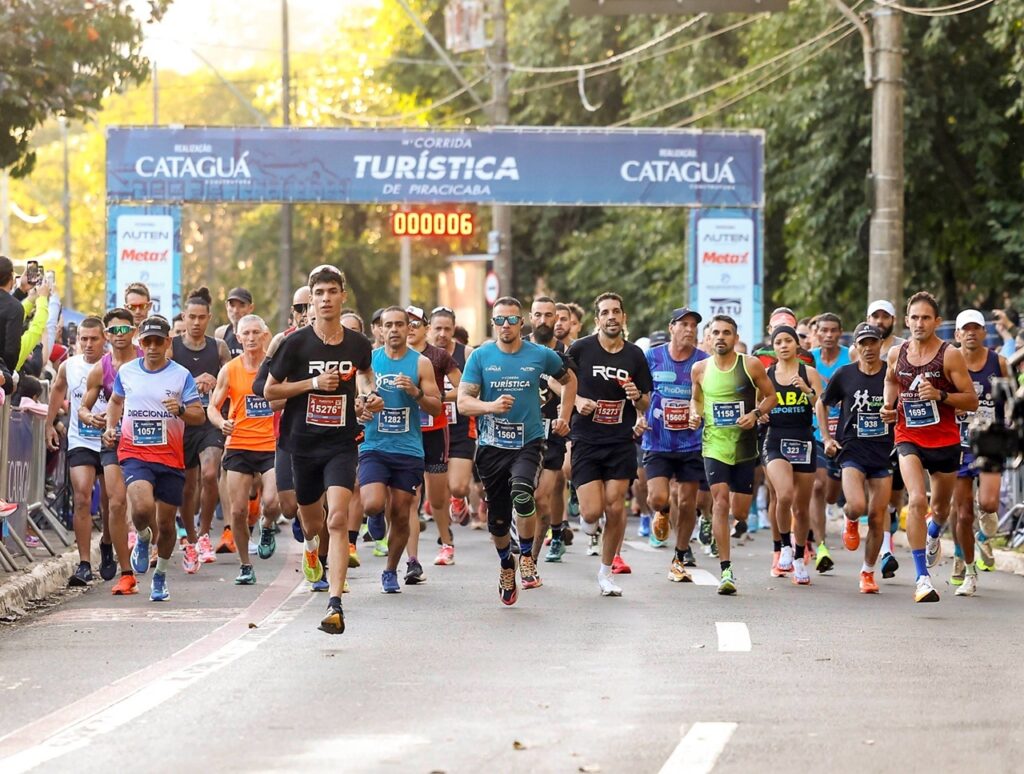A imagem mostra o início da 14ª Corrida Turística de Piracicaba, um evento de corrida de rua. Centenas de corredores estão em movimento, avançando pela rua em direção à câmera. Eles vestem roupas esportivas coloridas e números de identificação presos às camisetas. A energia e a concentração dos atletas são evidentes em suas expressões faciais. Ao fundo, há um grande arco azul com o nome do evento e o logotipo do patrocinador "Cataguá Construtora". Um relógio digital marca o tempo da corrida, indicando apenas seis segundos desde o início. A rua está cercada por árvores verdes e alguns espectadores podem ser vistos ao lado, assistindo e registrando o momento. A foto foi tirada em um dia claro, com luz natural que destaca as cores vibrantes da cena. A perspectiva frontal e a nitidez da imagem capturam a intensidade e o movimento do começo da prova.