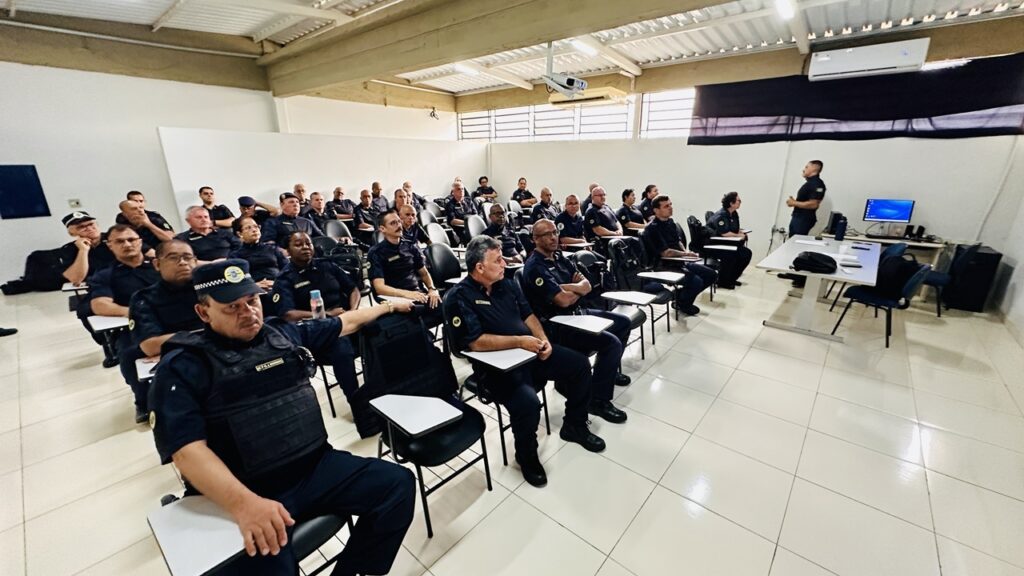A imagem mostra uma sala de aula onde um grupo grande de agentes da Guarda Municipal está participando de um treinamento ou palestra. Aproximadamente 30 a 35 pessoas, uniformizadas com roupas escuras e coletes táticos, estão sentadas em cadeiras com pequenas mesas acopladas, voltadas para a frente da sala. Um instrutor, também uniformizado, está em pé à frente, ao lado de uma mesa branca com um monitor de computador, aparentemente conduzindo a aula. A sala é ampla, com paredes brancas, piso claro e teto com estrutura metálica. Há um aparelho de ar-condicionado na parede, e um projetor está instalado no teto. A atmosfera é formal e profissional, indicando um momento de capacitação para os agentes.