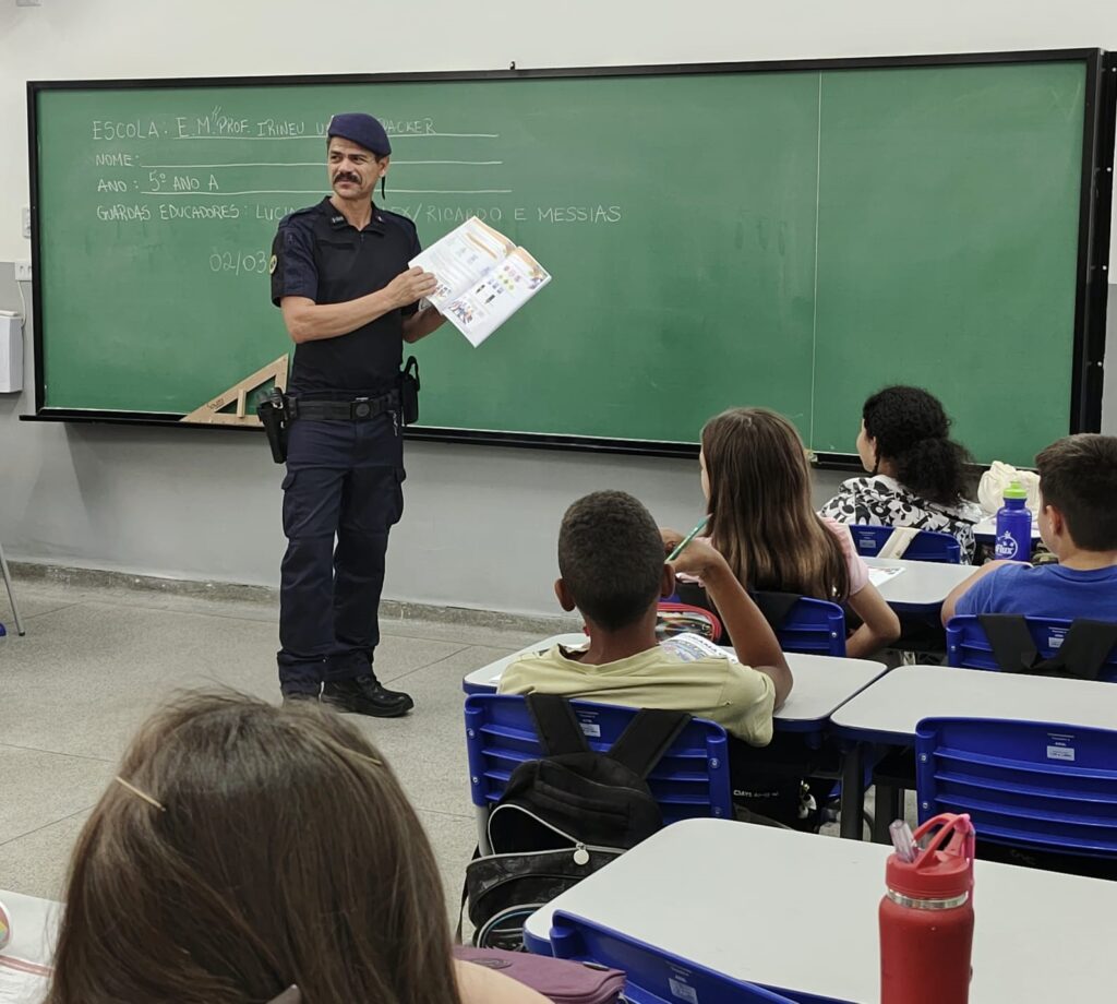 A imagem mostra uma sala de aula onde um homem adulto, provavelmente um guarda municipal, está em pé próximo ao quadro negro, falando para os alunos. Ele veste um uniforme azul escuro com um boné azul e está segurando um livro aberto. No quadro negro, escrito com giz branco, há informações sobre a escola, a turma (5º ano A) e os nomes dos guardas educadores: Lúcia, Ricardo e Messias. Alguns alunos estão sentados em carteiras, olhando para o homem. A sala tem paredes claras, cadeiras azuis e carteiras de madeira clara. O ambiente é iluminado e parece organizado, típico de uma escola.