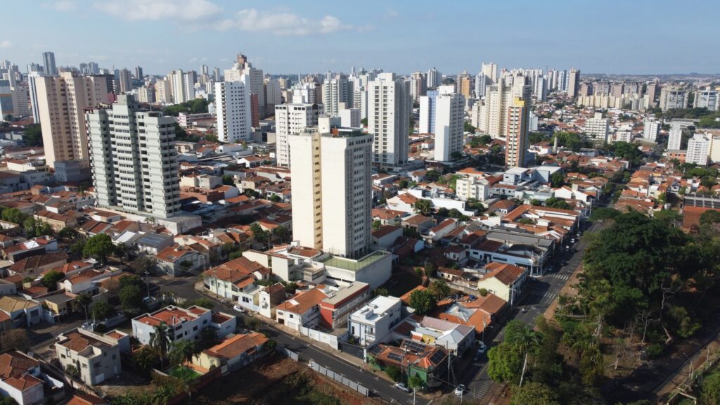 A imagem mostra uma vista aérea de uma grande área urbana, marcada pela diversidade arquitetônica. No centro e ao fundo, predominam edifícios altos e modernos, enquanto no primeiro plano aparecem casas menores com telhados vermelhos, ladeadas por ruas arborizadas. Essa composição evidencia o contraste entre bairros tradicionais de baixa densidade e o crescimento vertical característico das regiões metropolitanas.