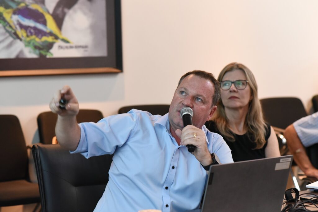 A imagem mostra um homem de meia-idade falando ao microfone, com expressão séria e gesticulando com a mão direita apontando para algo fora do quadro, à esquerda. Ele veste uma camisa social azul clara e usa aliança no dedo anelar esquerdo. Ao fundo, uma mulher loira, de óculos, observa atentamente, vestindo uma blusa preta. Eles estão sentados em cadeiras de couro pretas, provavelmente em uma sala de reunião ou escritório. Há um laptop aberto à frente do homem, sugerindo que ele pode estar apresentando ou discutindo algum assunto. Na parede ao fundo, há uma obra de arte com as cores da bandeira do Brasil (verde, amarelo e azul). A iluminação é clara e o foco principal está no homem, com o fundo levemente desfocado, destacando-o na cena.