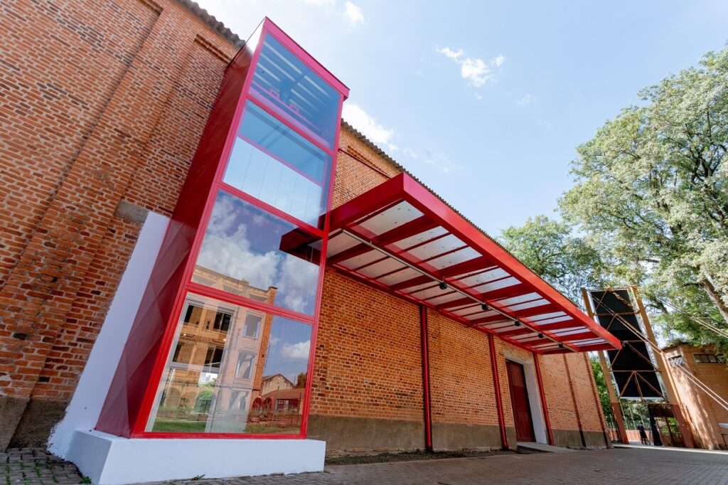 Na imagem aparece uma construção de tijolinhos e telhado vermelho. Ao lado aparece uma árvore verde clara com folhas brancas. Ao fundo aparece o céu azul e com pequena nuvem branca.