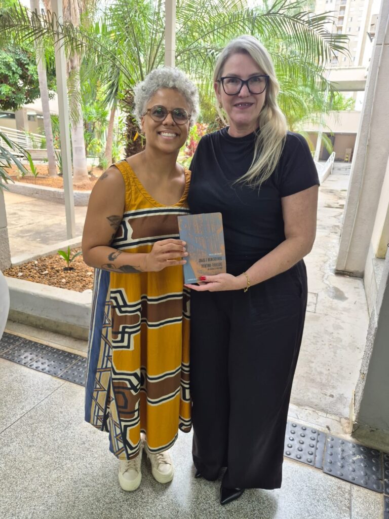 A imagem mostra duas mulheres em um ambiente externo, provavelmente um pátio ou área coberta, com vegetação tropical ao fundo, incluindo palmeiras. Elas estão posicionadas lado a lado, sorrindo para a câmera. A mulher à esquerda tem cabelo curto e grisalho, usa óculos redondos e um vestido amarelo com estampas geométricas em tons de marrom, branco e azul, além de ter tatuagens visíveis nos braços. A mulher à direita tem cabelo loiro e longo, usa óculos de armação preta e está vestida com uma camiseta preta e calça escura. Ambas seguram juntas um livro com capa azul e detalhes em laranja. No canto inferior direito da imagem, há um texto que parece ser o título de um trabalho ou livro: "MÁRCIA LIMA RIBEIRO - JOGOS E BRINCADEIRAS TRENTINO-TIROLEZAS NO ESTADO DE MINAS GERAIS - LUMENS DO SABER". O ambiente é iluminado por luz natural, e o chão é de pedra clara com faixas táteis para acessibilidade. A atmosfera parece ser amigável e profissional.