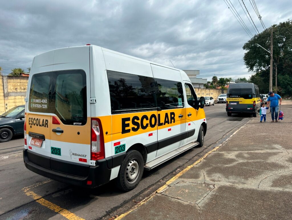 A imagem mostra uma van escolar branca da marca Renault, modelo Master, estacionada na rua. A van possui uma faixa amarela com a palavra "ESCOLAR" escrita em letras pretas tanto na lateral quanto na traseira do veículo. O veículo tem placa com o código "BAL-4441" e indica capacidade para 16 lugares. Ao fundo, é possível ver outra van escolar, de cor cinza escura com faixa amarela, e um homem segurando a mão de uma criança vestindo uniforme escolar azul e branco, caminhando em direção a essa segunda van. A rua está pavimentada, com uma calçada e uma faixa amarela na borda da via. O céu está nublado, sugerindo um dia com tempo fechado. Há também outros carros estacionados mais à frente na rua. A cena parece ser em uma área residencial ou próxima a uma escola, e os textos indicam que o local é provavelmente no Brasil, devido ao uso do português e à formatação dos telefones visíveis na imagem.