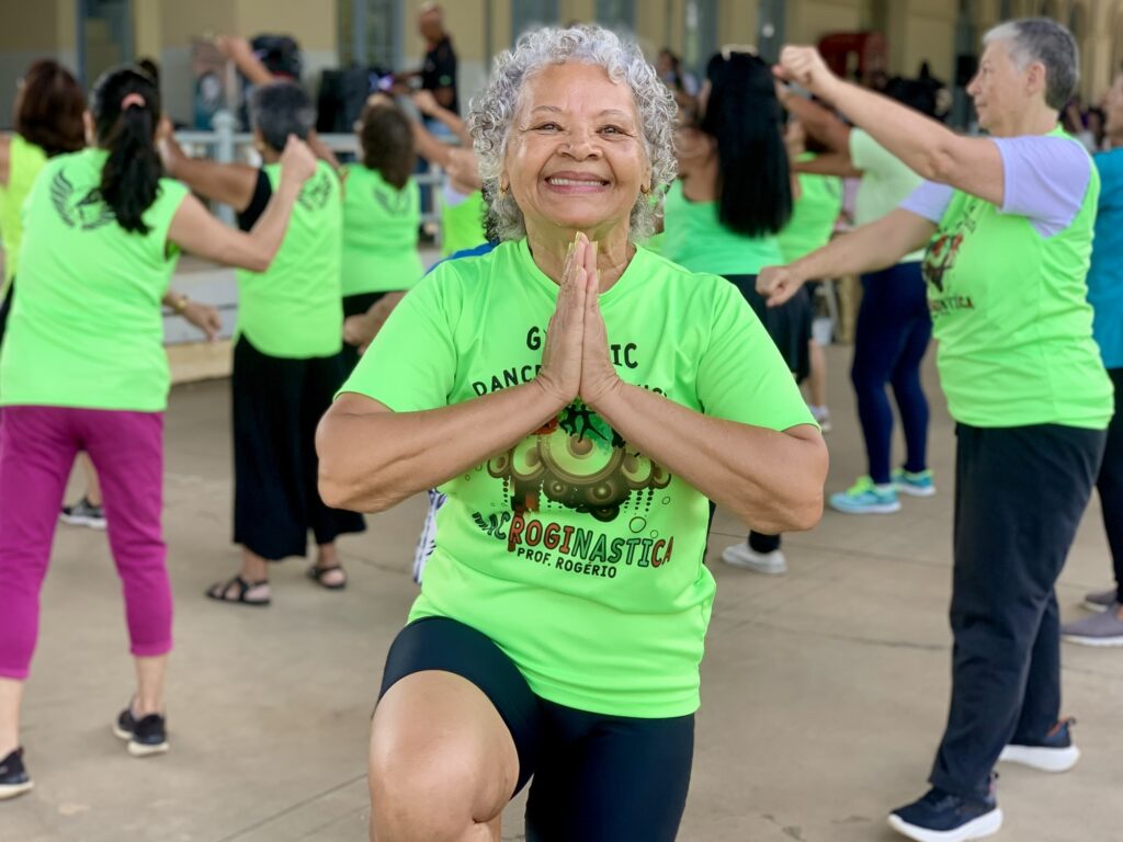 A imagem mostra uma mulher idosa sorridente, com cabelos grisalhos e encaracolados, participando de uma aula de ginástica ou dança em grupo. Ela está em destaque na frente, vestindo uma camiseta verde neon com os dizeres "GYM DANCE ROGINASTICA PROF. ROGÉRIO". A mulher está com as mãos unidas em posição de prece (Anjali Mudra), transmitindo serenidade e concentração. Ao fundo, outras pessoas, também vestindo camisetas verdes neon, participam da mesma atividade, formando um grupo que parece estar em movimento sincronizado. O ambiente é ao ar livre, possivelmente em uma praça ou área coberta com piso de concreto, com iluminação natural. A atmosfera geral é alegre e saudável, enfatizando a prática de exercícios físicos e o convívio social entre idosos.
