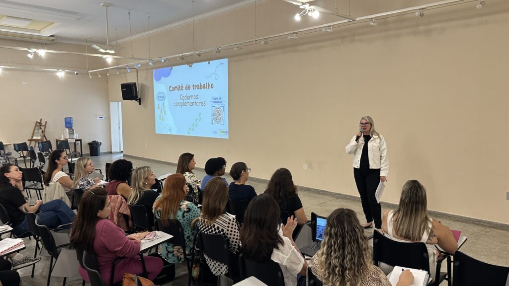 A imagem mostra uma sala de aula ou auditório onde está ocorrendo uma apresentação ou reunião. Na parte direita da imagem, uma mulher loira, vestindo uma jaqueta jeans branca e calça preta, está falando ao microfone, provavelmente conduzindo a sessão. À esquerda, um projetor exibe um slide com o título "Comitê de trabalho Cadernos complementares", a data "18/10/2023" e uma lista de presença com um QR code. À frente da palestrante, um grupo de cerca de 14 mulheres está sentado em cadeiras com apoio para escrever, prestando atenção à apresentação. Algumas estão com laptops, outras anotando ou ouvindo atentamente. A sala é ampla, com paredes claras e iluminação clara e uniforme, vinda de luminárias no teto. Ao fundo, há uma porta aberta, um bebedouro e alguns móveis. A atmosfera geral é de concentração e aprendizado, em um ambiente profissional e organizado.