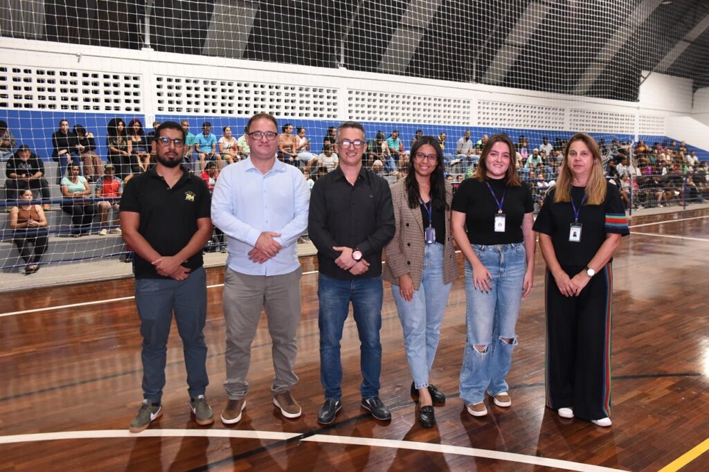 A imagem mostra um grupo de seis adultos posando juntos em uma quadra esportiva coberta. Há três homens à esquerda e três mulheres à direita, todos alinhados lado a lado. Os homens vestem roupas casuais, como camisa polo, camisa social e camiseta, combinadas com calças e tênis. As mulheres também estão vestidas de forma casual, usando blusas, blazer e jeans, algumas com crachás pendurados no pescoço. Ao fundo, há uma rede de proteção e várias pessoas sentadas em arquibancadas, assistindo ao evento. O chão da quadra é de madeira polida com marcações brancas e amarelas, e a iluminação é clara, típica de ginásios esportivos. A cena parece ser um momento de confraternização ou reconhecimento durante um evento esportivo comunitário ou escolar.