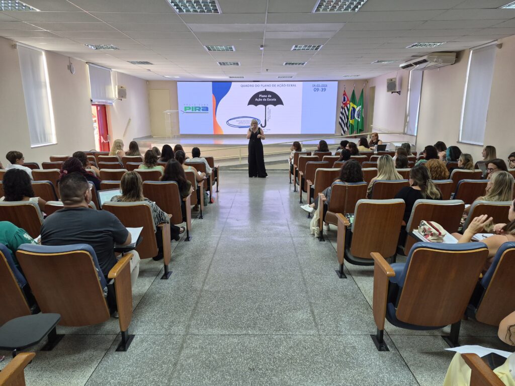 A imagem mostra uma mulher em um macacão preto apresentando em um auditório. Ela está de pé no centro do palco, de frente para o público, que está sentado em cadeiras de madeira com estofado azul, organizadas em fileiras ao longo de um corredor central. O ambiente é iluminado por luzes no teto, com paredes claras e piso cinza. Na parede atrás da apresentadora, há uma grande tela digital exibindo o título "QUADRO DO PLANO DE AÇÃO GERAL", a data "04-03-2024", o horário "09:39" e o texto "Plano de Ação da Escola", junto ao logo da "Prefeitura PIRA". Ao lado da tela, estão duas bandeiras, provavelmente do Brasil e do estado de São Paulo. O público é composto principalmente por mulheres, todas olhando para a apresentadora e a tela, indicando que estão assistindo a uma palestra ou apresentação oficial.