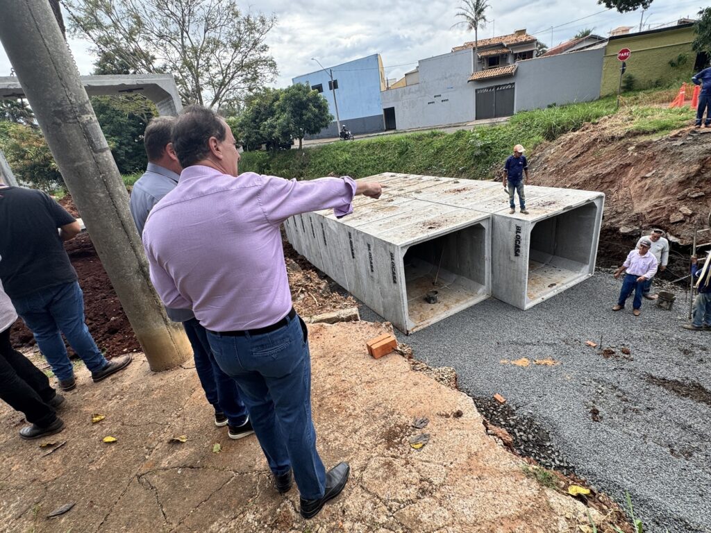 A imagem mostra um canteiro de obras ao ar livre, onde está sendo realizada a instalação de grandes blocos de concreto pré-moldados, conhecidos como caixas de concreto ou aduelas, provavelmente para um sistema de drenagem ou escoamento de água. Os blocos têm a marca "BLOCASA" estampada. No centro da imagem, há uma escavação profunda no solo, onde os blocos já estão posicionados sobre uma base de brita. Três operários estão dentro da escavação, em cima dos blocos, aparentemente trabalhando ou inspecionando a colocação. Na parte superior da escavação, fora dela, há três homens, sendo que um deles está apontando para os blocos no interior da vala. Eles parecem estar fazendo uma inspeção ou acompanhamento da obra. Dois deles vestem roupas sociais (camisa e calça), enquanto o outro está mais casual. Ao fundo, vê-se uma rua pavimentada com algumas casas e um poste com uma placa de trânsito vermelha com a palavra "PARE", indicando que a obra está localizada em uma área urbana, provavelmente no Brasil, devido à placa em português. O céu está nublado, indicando um dia com pouca luz natural. No geral, a imagem transmite a ideia de uma obra de infraestrutura, com foco na instalação de um sistema de drenagem subterrâneo, com supervisão técnica e trabalho em andamento.