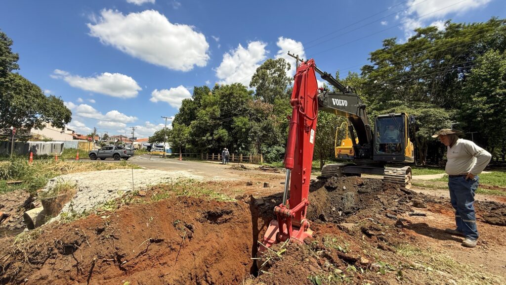 A imagem mostra uma cena de construção ao ar livre em um dia ensolarado. Em destaque, há uma escavadora grande da marca Volvo, com a estrutura principal amarela e o braço vermelho, posicionada sobre um terreno de terra vermelha. A escavadora está escavando uma vala profunda no solo. Ao lado da máquina, um homem está em pé, usando chapéu de aba larga, camisa de manga longa clara e calça jeans, observando o trabalho com as mãos na cintura. Outra pessoa está dentro da cabine da escavadora. Ao fundo, há uma estrada asfaltada com um caminhonete cinza estacionada e algumas pessoas caminhando. A vegetação verde e árvores cercam a área, e o céu está azul com nuvens brancas. Postes de energia e cabos elétricos aparecem na parte superior da imagem. A luz do sol forte cria sombras marcantes no chão e nas máquinas. A cena transmite uma sensação de atividade em um ambiente rural ou semiurbano.