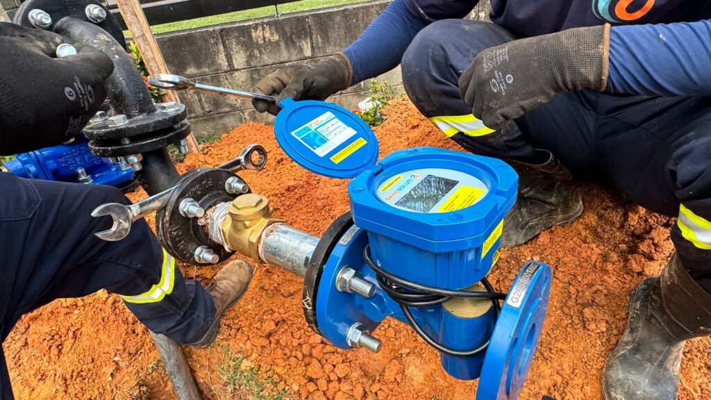 A imagem mostra dois trabalhadores realizando a instalação ou manutenção de um sistema de medição de água em uma tubulação subterrânea. Eles estão agachados, usando uniformes de trabalho azul escuro com faixas refletivas amarelas, luvas pretas e botas de segurança. O equipamento principal é um medidor de água azul, com a tampa aberta, onde se pode ver o mostrador do medidor. A tubulação é preta, com flanges e parafusos prateados, e há uma válvula de latão conectada. O solo ao redor é de terra vermelha, indicando que o local foi escavado para acessar a tubulação. Ao fundo, há uma parede de concreto e um pouco de grama verde. A imagem transmite uma cena típica de manutenção ou instalação de infraestrutura hidráulica em um ambiente externo. No medidor, aparecem inscrições como "SANE SOLUTI" e um número de série "G26SS0010002".