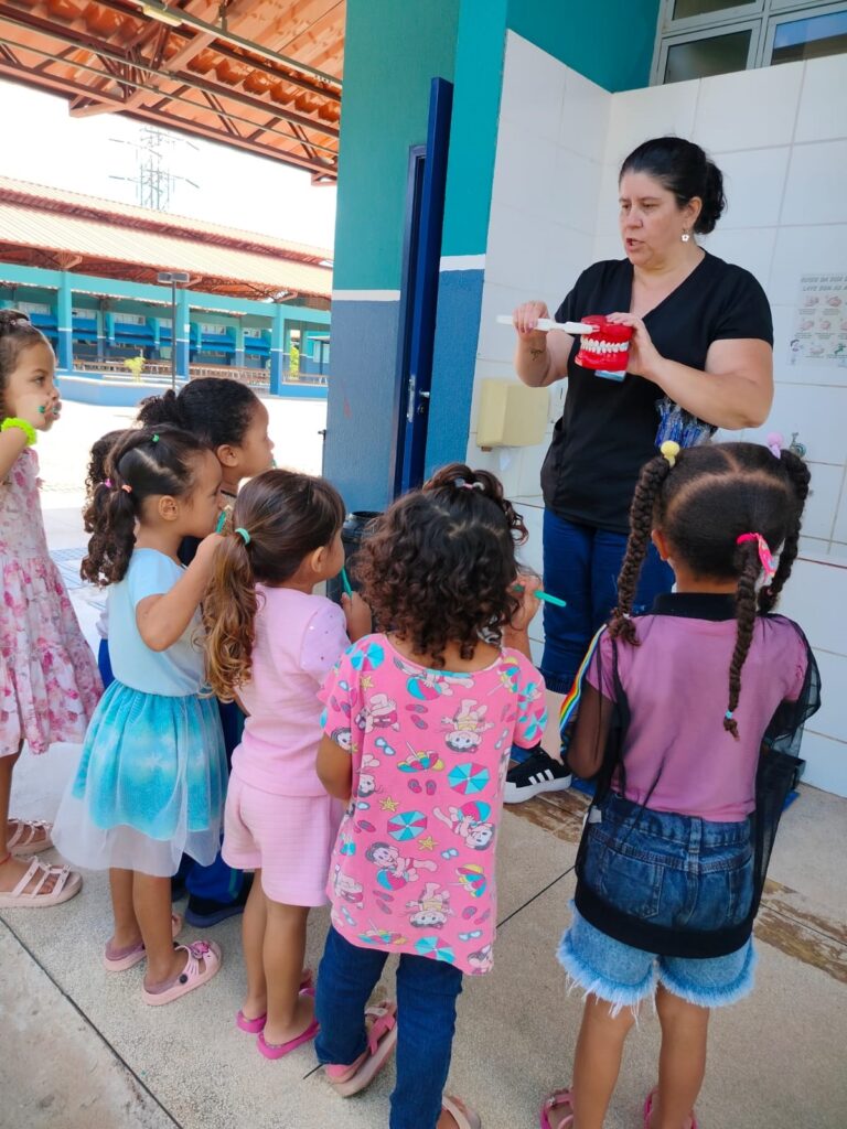 A imagem mostra uma cena ao ar livre em uma escola, onde uma mulher está ensinando um grupo de seis crianças pequenas sobre higiene bucal. A mulher, vestindo camiseta preta e calça azul, segura um modelo grande de dentes vermelhos e uma escova de dentes gigante branca, demonstrando como escovar os dentes corretamente. As crianças, todas meninas, estão em volta dela, observando atentamente e algumas segurando suas próprias escovas de dente. Elas usam roupas coloridas e têm diferentes estilos de cabelo, como tranças e cabelos cacheados. Ao fundo, há uma parede branca com azulejos e um cartaz pequeno com instruções de higiene, além de uma estrutura com teto de telhas. A luz natural do sol ilumina a cena, que transmite um momento educativo e envolvente sobre cuidados com a saúde bucal.