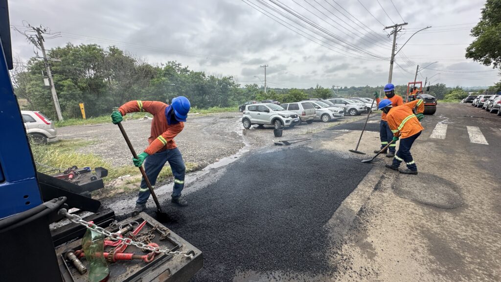 A imagem mostra um grupo de trabalhadores de construção rodoviária realizando a pavimentação de um trecho de asfalto. Vestindo camisas laranja de segurança, capacetes azuis, luvas verdes e faixas refletivas, eles estão concentrados em espalhar e nivelar o material recém-aplicado: um deles utiliza uma pá para retirar o asfalto da máquina, enquanto os demais o distribuem e alisam com ferramentas próprias. Ao fundo, vê-se um estacionamento com carros parados, postes com fiação elétrica e um céu nublado, compondo o cenário de uma atividade essencial de manutenção de infraestrutura urbana.