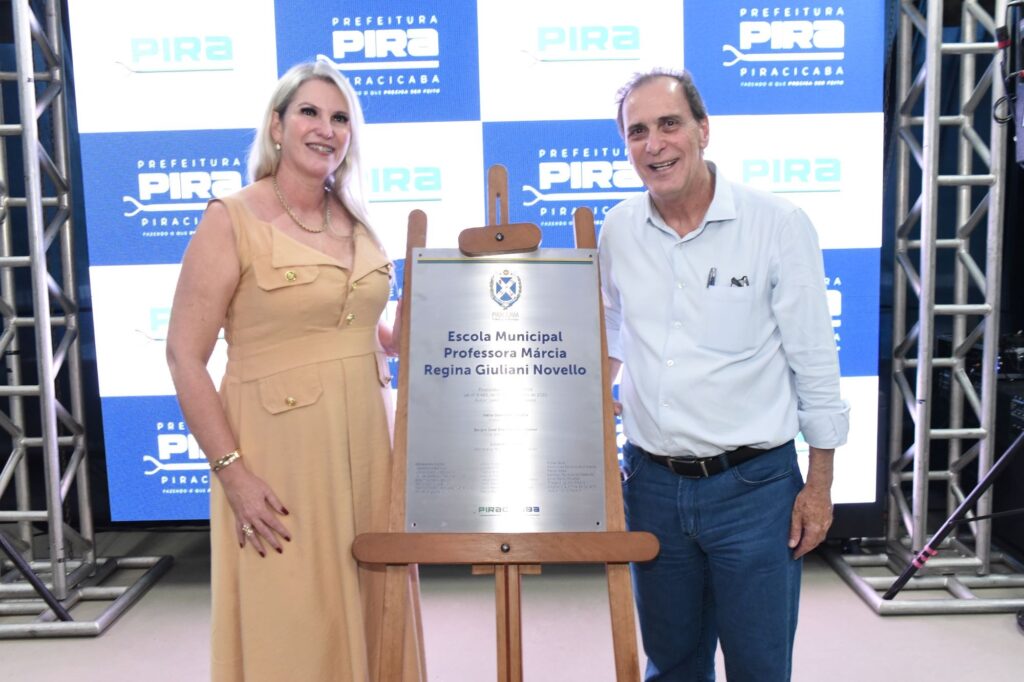 A imagem mostra duas pessoas, um homem e uma mulher, posando ao lado de uma placa comemorativa em um cavalete de madeira. A mulher está à esquerda, usando um vestido sem mangas na cor mostarda, com botões dourados, e usa um colar e pulseira dourados. Ela sorri para a câmera. O homem está à direita, vestindo uma camisa azul clara de mangas compridas e calça jeans, também sorrindo. Entre eles está uma placa prateada com o brasão da cidade de Piracicaba e texto em português que indica a inauguração da "Escola Municipal Professora Márcia Regina Giuliani Novello" em 2024. Ao fundo, há um painel azul e branco com o logotipo da Prefeitura de Piracicaba repetido. A cena parece ser uma cerimônia oficial de inauguração ou homenagem, com iluminação clara e ambiente interno.