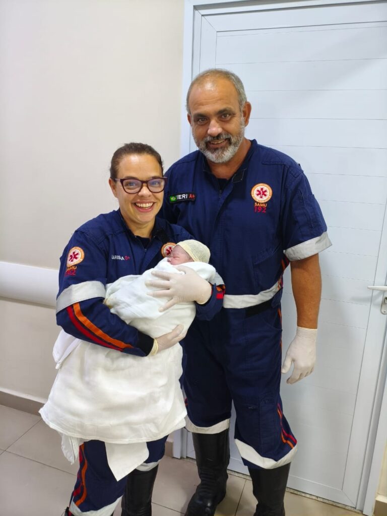 A imagem mostra dois profissionais de saúde do SAMU (Serviço de Atendimento Móvel de Urgência) sorrindo enquanto seguram um recém-nascido envolto em um cobertor branco. Ambos vestem uniformes azuis escuros com o símbolo da Estrela da Vida e a inscrição "SAMU 192". A mulher, com óculos e cabelo preso, segura o bebê com cuidado e alegria, enquanto o homem, com barba grisalha, está ao lado dela, também sorrindo. O bebê usa um pequeno gorro amarelo. O ambiente parece ser um corredor hospitalar, com paredes claras e uma porta branca ao fundo. A cena transmite um momento de felicidade e alívio, possivelmente após um atendimento de emergência bem-sucedido.