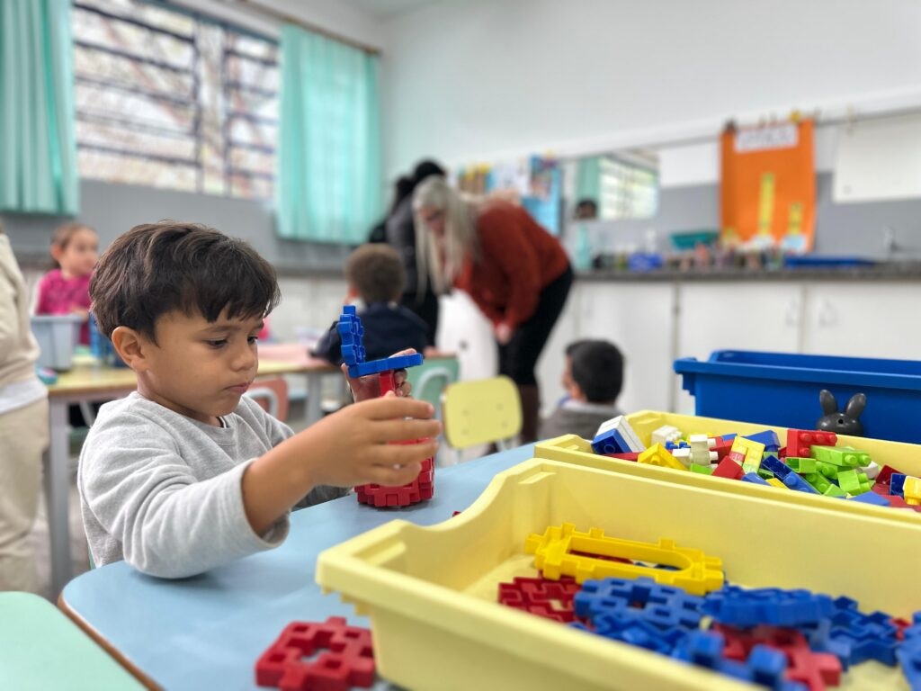 A imagem mostra um menino pequeno, com cabelo escuro e camiseta cinza de manga comprida, sentado em uma mesa azul. Ele está brincando com blocos de montar coloridos, segurando uma estrutura feita com blocos vermelhos e azuis. Na frente dele, há caixas amarelas cheias desses blocos. Ao fundo, é possível ver um ambiente de sala de aula, com outras crianças e duas adultas, uma delas está inclinada para interagir com uma das crianças. A sala é iluminada por luz natural que entra por uma janela com cortinas azul claro. O cenário transmite um ambiente alegre e educativo, típico de uma pré-escola ou creche.