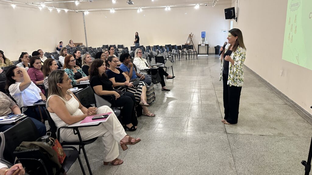 A imagem mostra uma palestra ou workshop acontecendo em uma sala ampla e bem iluminada. No lado direito, uma mulher está de pé, segurando um microfone, provavelmente a palestrante, vestindo uma blusa preta com um casaquinho branco estampado com limões. À esquerda, várias mulheres estão sentadas em cadeiras pretas com apoio para anotações, prestando atenção à apresentação. Algumas delas estão anotando, outras apenas observam atentamente. Ao fundo, há uma tela de projeção exibindo um slide com a data "09/03/2024" e imagens de lâmpadas, sugerindo um tema relacionado a ideias ou inovação. A sala tem paredes claras e um piso brilhante, com iluminação proveniente de luminárias no teto. No ambiente, também é possível ver um bebedouro e alto-falantes fixados na parede. A atmosfera parece profissional e focada no aprendizado ou desenvolvimento.