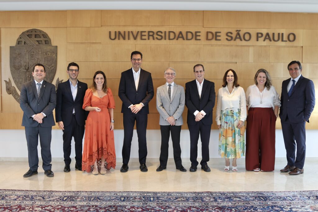 A imagem mostra um grupo de nove pessoas posando para uma foto em frente a uma parede de madeira clara. Na parede, está escrito "UNIVERSIDADE DE SÃO PAULO" em letras grandes e escuras, ao lado de um brasão metálico. O grupo é composto por seis homens e três mulheres, todos vestidos de forma profissional, com ternos, vestidos e roupas sociais. Eles estão alinhados, sorrindo e olhando diretamente para a câmera. O ambiente parece ser uma área interna da universidade, com piso claro e uma pequena parte de um tapete azul visível na parte inferior da imagem. A iluminação é clara e uniforme, dando a impressão de uma foto oficial ou institucional.