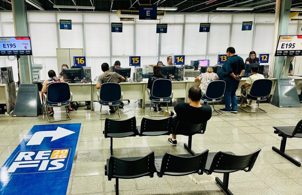 A imagem mostra o interior de um ambiente administrativo, provavelmente uma unidade de atendimento público da Prefeitura de Piracicaba, no Brasil. O local é bem iluminado com luzes fluorescentes e tem um visual moderno e organizado, com paredes brancas e piso cinza. Na parte de trás da sala, há um balcão longo dividido em vários postos de atendimento, numerados de 12 a 16, todos identificados com a placa "ATENDIMENTO DÍVIDA ATIVA". Cada posto possui computadores, cadeiras azuis para os atendentes e equipamentos de escritório. Diversos servidores estão sentados atendendo os cidadãos. Na frente, há uma área de espera com bancos pretos onde algumas pessoas aguardam seu atendimento. No chão, há um adesivo grande com a inscrição "REFIS 2026", que indica um programa de regularização fiscal. Acima do balcão, um painel digital exibe números de senha, como "E195", e indica o local do atendimento, por exemplo "Mesa 6". Um letreiro azul com a palavra "SENHAS E" também está visível, sugerindo o local para retirada de senhas. No geral, o ambiente transmite organização e eficiência no atendimento ao público para questões relacionadas a dívidas ativas e regularização fiscal.