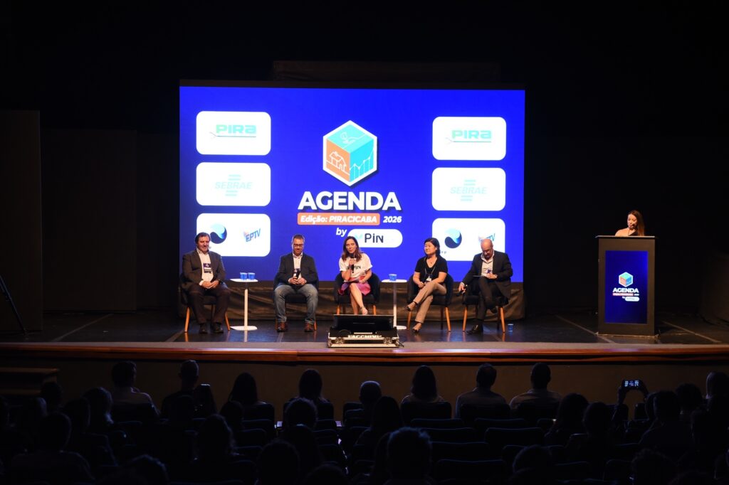 A imagem mostra um evento de painel de discussão realizado em um auditório escuro. No palco, cinco pessoas estão sentadas em cadeiras, três homens e duas mulheres, sendo que uma das mulheres está falando ao microfone, vestindo uma camiseta branca e saia rosa. As outras pessoas no palco estão vestidas de forma casual e elegante, com blazers e calças. À direita, uma mulher está em pé atrás de um púlpito com a marca do evento. Ao fundo, uma grande tela azul exibe o logo do evento, que é um cubo 3D com ícones que representam habitação, natureza e crescimento, além do título "AGENDA Edição: PIRACICABA 2026". Também aparecem os logos dos patrocinadores ou apoiadores, como PIRA, SEBRAE e EPTV. O público está em primeiro plano, mas aparece em silhueta devido à iluminação focada no palco. A ambientação sugere um evento formal, possivelmente uma conferência ou fórum, com discussões sobre o futuro da cidade de Piracicaba até o ano de 2026. A iluminação destaca os participantes do painel, enquanto o restante do auditório permanece escuro. Pequenas mesas com copos d'água estão posicionadas entre as cadeiras dos debatedores.
