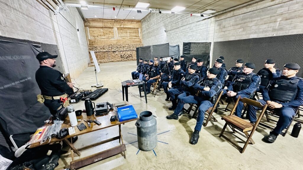 A imagem mostra uma sessão de treinamento em um ambiente interno, provavelmente uma sala de aula ou estande de tiro adaptado para instrução. Ao centro e à esquerda, um instrutor vestido com uniforme tático e boné preto está em pé atrás de uma mesa de madeira, que contém diversos objetos, como um laptop, estojos, garrafas e materiais de treinamento. À direita, um grupo de cerca de 18 pessoas, todas vestindo uniformes táticos azul-escuro e boinas pretas, está sentado em cadeiras dobráveis de madeira, prestando atenção ao instrutor. Ao fundo, há uma parede de concreto com uma estrutura de madeira que parece ser o alvo de um estande de tiro. No canto esquerdo da imagem, uma faixa preta com o texto "COMUNICAÇÃO CORPORAL" está parcialmente visível, indicando que o tema da aula pode estar relacionado à linguagem corporal ou comunicação não verbal. O ambiente é iluminado por luzes artificiais, com um tom profissional e sério, típico de treinamentos militares ou policiais. No chão, em primeiro plano, há um grande cilindro metálico, possivelmente parte do equipamento do local. A imagem transmite uma atmosfera de concentração e aprendizado.