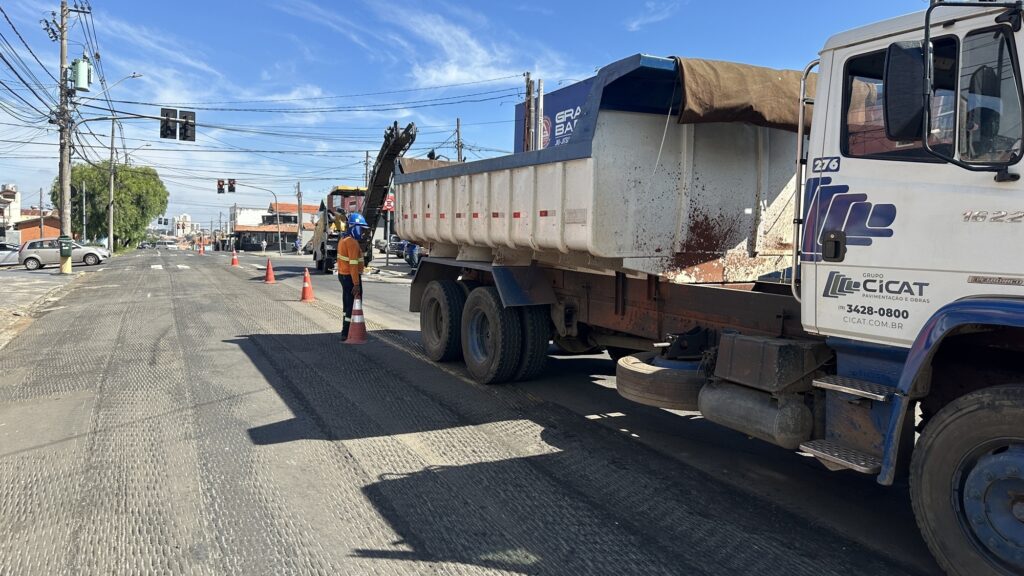 A imagem mostra uma cena de obras em uma rua durante o dia. No centro, há um caminhão basculante branco e azul da empresa "GRUPO CICAT PAVIMENTAÇÃO E OBRAS", com os números 276 e 1622 visíveis na cabine. O caminhão está parado sobre uma superfície de asfalto fresado, que apresenta marcas típicas do processo de fresagem. Ao lado do caminhão, um trabalhador usando colete laranja, capacete azul e máscara está segurando um cone de sinalização laranja. Ao fundo, é possível ver uma máquina fresadora em operação, descarregando material em outro veículo. A cena está delimitada por vários cones de sinalização, indicando a área de trabalho. O céu está claro com poucas nuvens, e na lateral esquerda da imagem há um poste de energia com transformador e fios elétricos, além de um carro prata estacionado próximo a uma lixeira verde. A foto foi tirada de um ângulo baixo, evidenciando a dimensão do caminhão e da máquina.