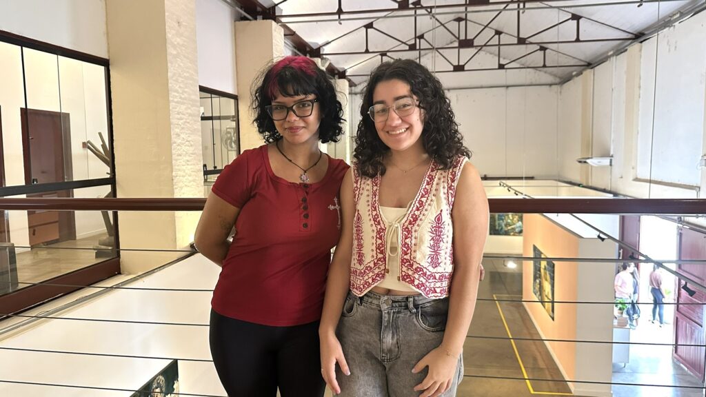 A imagem mostra duas mulheres jovens, posicionadas lado a lado, apoiadas em um corrimão de metal e madeira, em um espaço interno que parece ser uma galeria de arte ou museu. Elas estão sorrindo e olhando para a câmera. A mulher à esquerda tem cabelo curto com mechas rosas, usa óculos, camiseta vermelha e calça preta. A mulher à direita tem cabelo longo e cacheado, também usa óculos, uma blusa branca com um colete vermelho bordado e calça cinza. O ambiente é amplo, com teto alto e estruturas metálicas aparentes, além de quadros pendurados nas paredes ao fundo. A iluminação é natural, entrando pela direita da imagem, e destaca as cores das roupas das mulheres, criando um contraste com as paredes claras e o piso escuro da galeria. A composição transmite uma atmosfera acolhedora e cultural.
