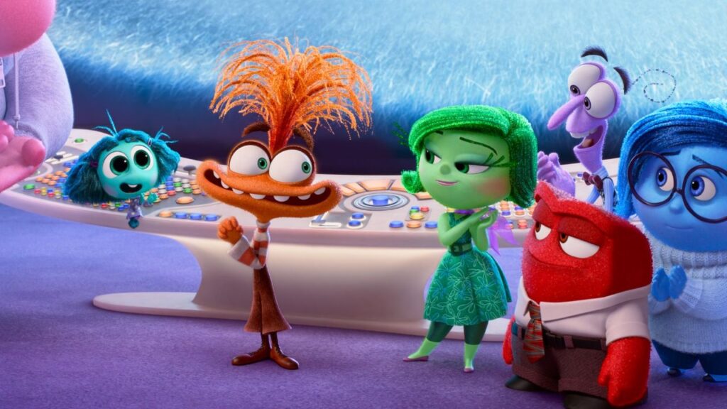 A imagem é uma cena animada de alta qualidade do filme "Divertida Mente 2", mostrando várias emoções personificadas em frente a um console de controle futurista. No centro está a personagem Ansiedade, com cabelos laranja arrepiados, olhos arregalados e uma expressão animada, vestindo uma camisa listrada e calças marrons. À esquerda dela está Inveja, uma pequena personagem verde-azulada com olhos grandes. À direita de Ansiedade está Nojinho, uma personagem verde com cabelo curto e vestido floral, com uma expressão levemente cética. Em primeiro plano, à direita, está Raiva, uma personagem vermelha, quadrada, com expressão carrancuda, vestindo camisa branca e gravata. Ao fundo, à direita, aparecem Medo (roxo) e Tristeza (azul, com óculos). O ambiente é a sala de controle da "Sede", com um console branco curvo cheio de botões coloridos e luminosos. A iluminação é suave e clara, destacando as texturas variadas dos personagens, como o cabelo desgrenhado de Ansiedade e a pele áspera de Raiva. A cena transmite uma atmosfera energética e um pouco caótica, refletindo a introdução das novas emoções.