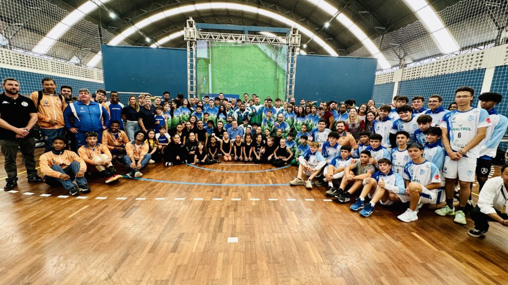 A imagem mostra uma grande equipe esportiva reunida em um ginásio coberto, posando para uma foto em grupo. O grupo é composto principalmente por jovens atletas, vestidos com uniformes de diferentes cores: laranja e preto à esquerda, verde e branco ao centro, e azul claro e branco à direita. Alguns estão sentados no chão em primeiro plano, enquanto a maioria está em pé, organizados em filas que seguem a linha de três pontos da quadra de basquete. O piso é de madeira, típico de quadras esportivas, e o ambiente é iluminado por luzes no teto arqueado do ginásio. Ao fundo, há uma grande tela exibindo uma imagem de campo verde, com cortinas azuis nas laterais e uma estrutura metálica que a sustenta. No alto, é possível ver uma rede de proteção. A atmosfera sugere um momento formal, possivelmente o encerramento de um evento esportivo ou um registro oficial da equipe. Palavras extraídas da imagem indicam nomes e temas relacionados ao esporte, como "ESPORTE" e "MANUTROL".