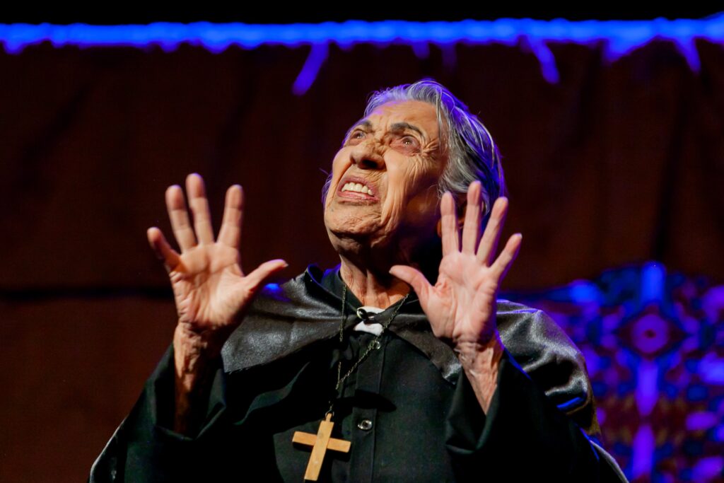 A imagem mostra uma mulher idosa em uma pose dramática e teatral. Ela está olhando para cima com uma expressão intensa, que pode ser de agonia, súplica ou desespero. Seu rosto está marcado por rugas profundas, e sua boca está ligeiramente aberta, mostrando os dentes. As mãos estão levantadas e abertas ao nível dos ombros, como se estivesse implorando ou expressando um forte sentimento. Ela veste uma roupa preta que lembra um hábito clerical, com uma pequena gola branca visível e uma capa curta preta sobre os ombros. Um grande crucifixo de madeira está pendurado no pescoço dela por uma corrente. A iluminação é dramática, destacando as texturas da pele envelhecida e as dobras da roupa, contra um fundo escuro em tons marrons e com um leve padrão azul visível à direita. Acima da cabeça dela, há uma linha de luz azul vibrante que parece fazer parte da cenografia ou iluminação cênica. A perspectiva da foto é levemente de baixo para cima, o que realça o olhar voltado para cima e a intensidade da cena, sugerindo que a imagem foi capturada em um momento emocional forte, possivelmente durante uma apresentação teatral ou cena de filme.