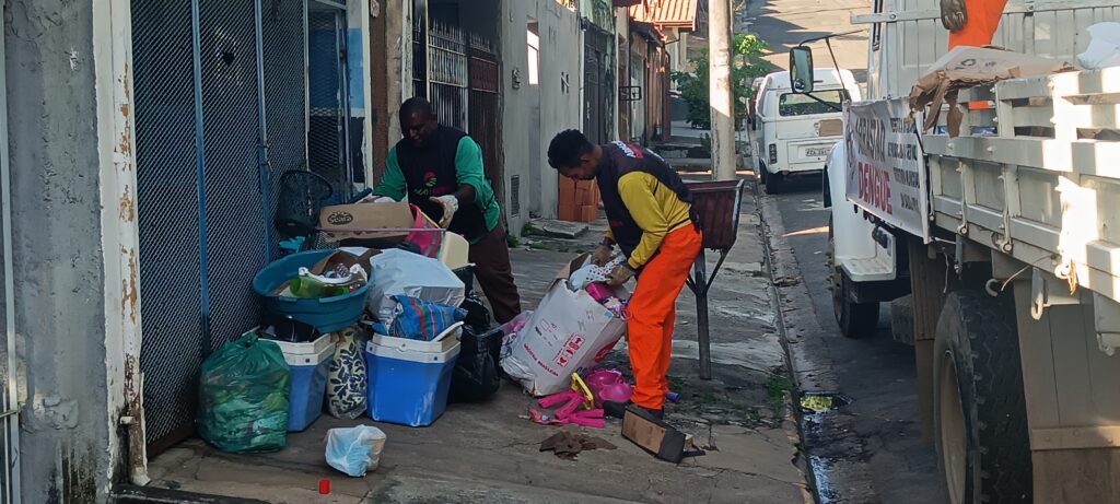 A imagem mostra dois trabalhadores de limpeza ou agentes de saúde pública recolhendo lixo e objetos descartados em uma calçada de uma rua urbana. Eles estão próximos a um muro com portas metálicas e a um caminhão branco estacionado ao lado, que tem uma placa com a palavra "DENGUE" em destaque, indicando uma ação de combate à dengue, provavelmente removendo possíveis criadouros do mosquito. Um dos homens veste calça laranja e camiseta amarela, e está agachado mexendo em um saco branco grande, enquanto o outro, com calça marrom e colete escuro, observa ou ajuda na coleta. No chão há várias caixas de papelão, um saco verde e dois recipientes plásticos azuis, além de lixo espalhado. A cena ocorre em um dia ensolarado, com sombras marcadas. Ao fundo, é possível ver um veículo branco com placa "FEA 2817" e alguns prédios antigos, sugerindo um bairro residencial ou comercial. A ação retratada é uma medida de saúde pública para eliminar focos do mosquito da dengue, reforçando a importância da limpeza urbana para a prevenção de doenças.