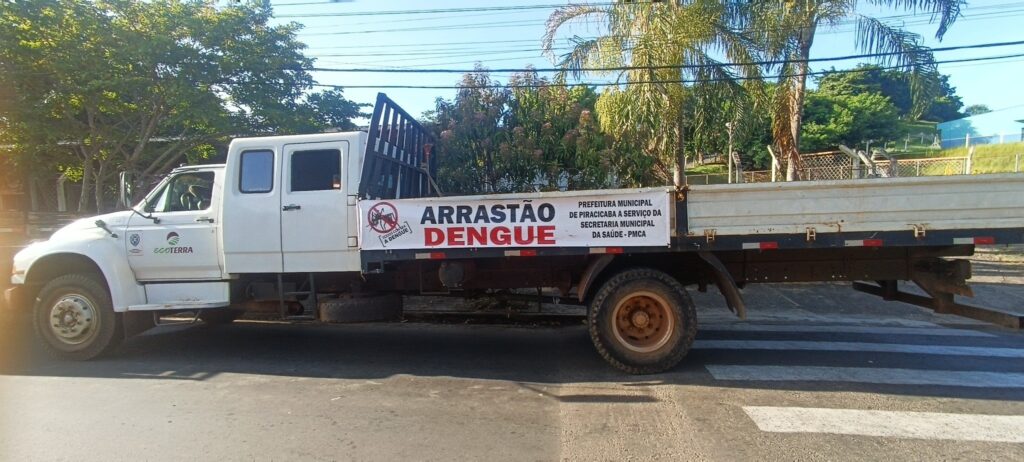A imagem mostra uma caminhonete branca com cabine dupla estacionada em uma rua asfaltada, próxima a uma faixa de pedestres. A caminhonete possui uma carroceria aberta, onde está fixada uma faixa branca grande com a inscrição "ARRASTÃO DENGUE" em letras grandes, sendo "ARRASTÃO" em preto e "DENGUE" em vermelho. Ao lado do texto, há um símbolo de mosquito dentro de um círculo vermelho com uma linha diagonal, indicando proibição. A faixa também traz o texto "TODOS CONTRA A DENGUE" e a identificação da Prefeitura Municipal de Piracicaba, informando que o veículo está a serviço da Secretaria Municipal da Saúde. A cabine da caminhonete tem o logo "ecoterra". Ao fundo, é possível ver árvores verdes e palmeiras sob um céu azul claro, com fios elétricos visíveis no alto da imagem. A cena está bem iluminada pelo sol, com sombras nítidas no chão.