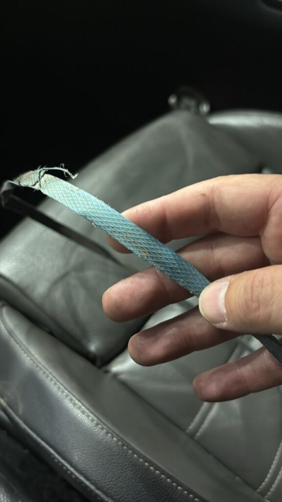A imagem mostra uma mão segurando uma correia azul, que está visivelmente desgastada e danificada, com fios soltos e partes desfiadas. A correia possui um padrão quadriculado em relevo. A mão aparenta estar suja, com marcas de sujeira ou graxa, sugerindo que a pessoa está trabalhando com alguma manutenção mecânica. Ao fundo, vê-se o banco de um carro, indicando que a correia provavelmente é parte do sistema do veículo. A foto foi tirada de perto, focando na correia e na mão, com o fundo desfocado.