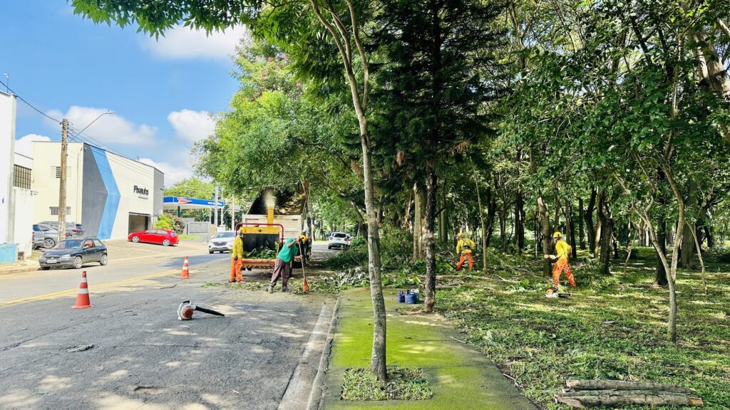 A imagem mostra uma equipe de manutenção urbana trabalhando na poda e limpeza de árvores ao longo de uma rua ensolarada. À esquerda, há uma estrada asfaltada onde está estacionado um caminhão branco equipado com um triturador de galhos. Dois trabalhadores com roupas de segurança — um de camiseta verde e outro com colete laranja — estão recolhendo os detritos. À direita, entre as árvores, dois outros trabalhadores em camiseta amarela e calça laranja utilizam motosserras para cortar galhos. No primeiro plano, há um soprador de folhas sobre o chão e cones laranjas indicam a área de trabalho. Ao fundo, observa-se um prédio comercial com a marca "Fixauto" e um posto de combustível, sob um céu azul com algumas nuvens. A cena transmite uma ação organizada de manutenção urbana em um ambiente com clima tropical ou subtropical.
