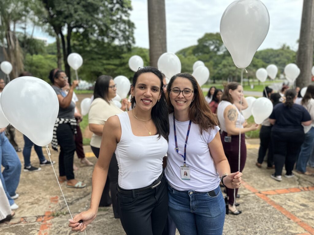 A imagem mostra duas mulheres sorrindo, em um ambiente ao ar livre, possivelmente em um parque ou praça com muitas árvores e palmeiras ao fundo. Ambas seguram balões brancos presos a fitas. A mulher à esquerda tem cabelo longo e escuro, veste uma regata branca, calça preta e um cinto preto, além de usar um colar dourado. A mulher à direita usa óculos, camiseta branca, calça jeans azul e um crachá pendurado no pescoço com um cordão azul. Ao fundo, outras pessoas também seguram balões brancos, sugerindo que pode ser um evento ou celebração. O céu está nublado, proporcionando uma luz suave e difusa na cena. O ambiente é agradável e transmite uma atmosfera de confraternização e alegria.