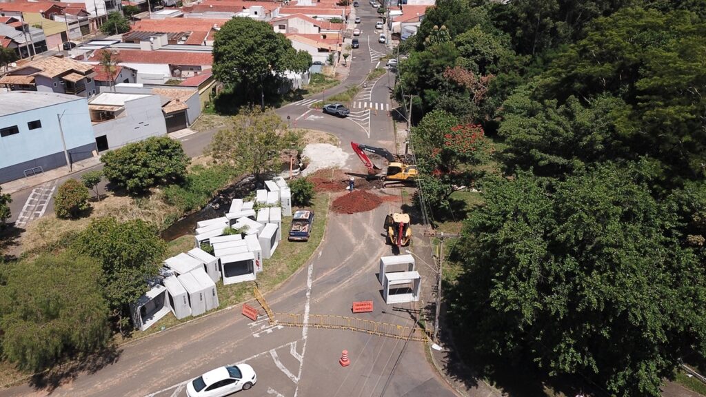A imagem mostra uma obra de construção ou manutenção em uma estrada localizada em uma área residencial. No centro da imagem, há um grande maquinário amarelo e vermelho da marca Volvo, possivelmente uma escavadeira, trabalhando próximo a uma pilha de terra vermelha. Ao redor, há vários blocos grandes de concreto, que parecem ser tubos ou estruturas para drenagem, espalhados sobre a grama e ao longo da via. A estrada está bloqueada com barreiras amarelas e cones laranja, e há uma placa vermelha com a inscrição "TRÂNSITO IMPEDIDO", indicando que o tráfego está interrompido no local. Próximo à barreira, há um carro branco parado. À esquerda da estrada, há um bairro residencial com casas de diferentes cores e telhados vermelhos, enquanto à direita há uma área de vegetação densa e verde. O ambiente está iluminado por luz solar forte, sugerindo que a foto foi tirada durante o dia, provavelmente ao meio-dia. A perspectiva é aérea, possivelmente capturada por um drone, mostrando uma visão ampla da obra e da área ao redor.