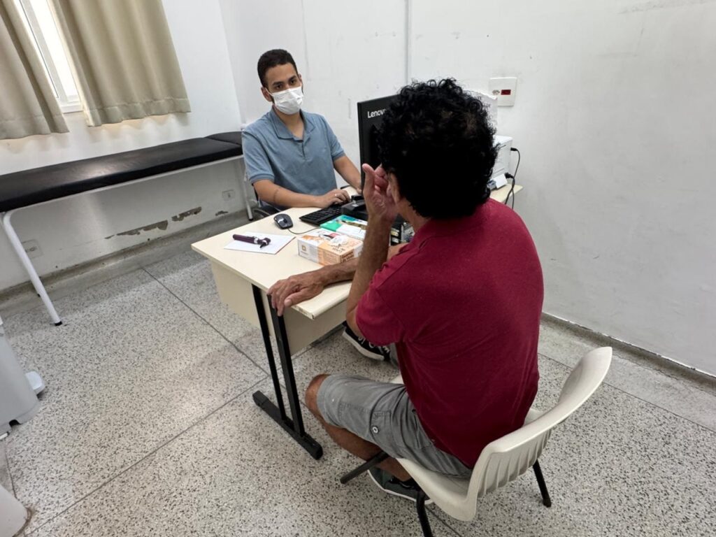 A imagem mostra uma consulta médica em um consultório bem iluminado. À esquerda, um profissional de saúde, provavelmente um médico, está sentado atrás de uma mesa, vestindo uma camisa polo azul clara e uma máscara facial branca. Ele olha atentamente para o paciente, com as mãos próximas ao teclado do computador. À direita, um homem mais velho, visto de costas, usa uma camiseta vinho e shorts cinza, gesticulando com a mão direita enquanto conversa com o médico. Sobre a mesa há um monitor Lenovo, teclado, mouse, uma caixa de luvas de látex e um pequeno instrumento médico sobre um papel. O ambiente é limpo, com paredes brancas, piso de tom cinza e uma maca preta ao lado da parede, sob uma janela com cortinas bege. A cena transmite uma interação profissional e atenciosa entre médico e paciente.