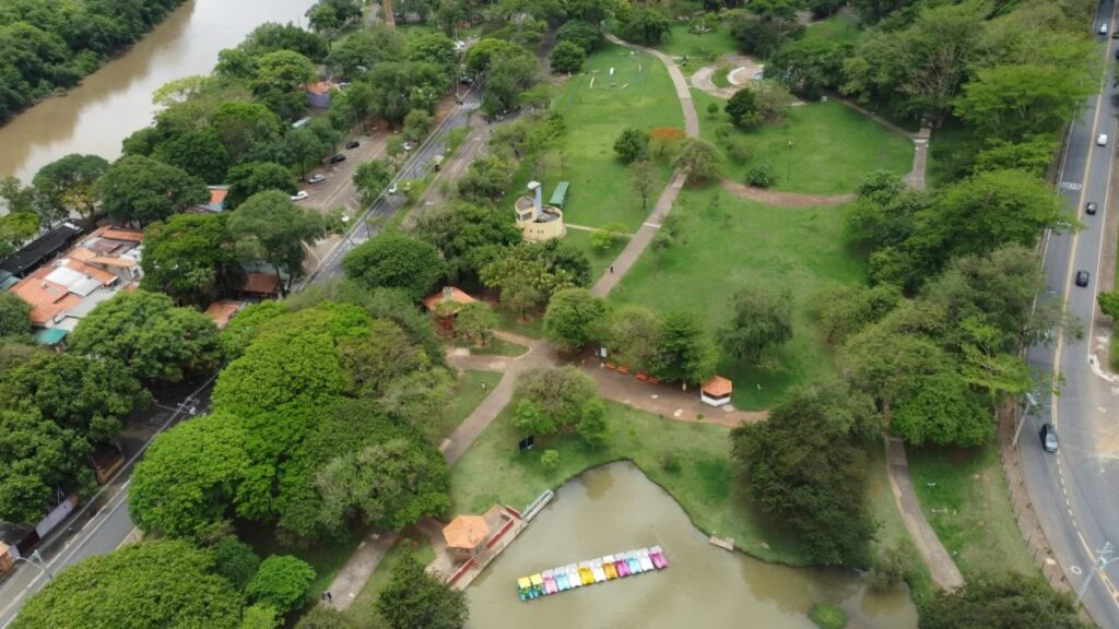 A imagem mostra uma vista aérea de um parque urbano com muita área verde, árvores e gramados bem cuidados. Há diversas trilhas e caminhos que cortam o parque, possibilitando o passeio das pessoas. Próximo ao centro da imagem, há um pequeno lago com pedalinhos coloridos (azul, amarelo, rosa e branco) alinhados na margem. Também é possível ver algumas construções pequenas dentro do parque, como um quiosque e uma estrutura circular amarela com uma torre. Ao redor do parque, há ruas com carros estacionados e em movimento, além de uma área com casas de telhados vermelhos e um rio de água barrenta. A iluminação está uniforme, sugerindo um dia nublado ou com pouca sombra direta. Na via ao lado do parque, há uma pintura no asfalto com a palavra "ATENÇÃO", indicando um alerta para os motoristas. No geral, a imagem transmite uma sensação de tranquilidade e lazer em meio à cidade, mostrando um espaço verde bem cuidado e utilizado para atividades recreativas.