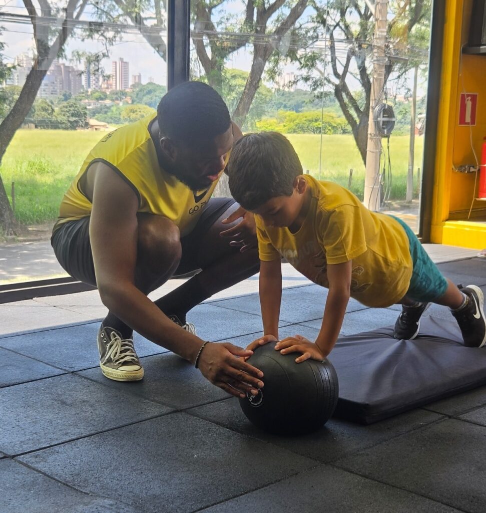 A imagem mostra um momento de treino em uma academia moderna, com grandes janelas que deixam entrar muita luz natural. Um homem, que parece ser um treinador, está ajudando um menino a fazer um exercício com uma bola medicinal preta. O menino está apoiado com as mãos na bola, em uma posição que lembra uma prancha, enquanto o treinador está ao lado, orientando e apoiando para garantir que ele faça o movimento corretamente. Ambos vestem roupas esportivas com tons de amarelo — o treinador usa uma regata amarela e o menino uma camiseta amarela com shorts azul-esverdeado estampado. O ambiente é amplo, com piso preto e equipamentos de academia ao redor. Ao fundo, é possível ver uma paisagem verde e o horizonte da cidade através das janelas. A cena transmite uma atmosfera de cuidado, aprendizado e incentivo, destacando a interação entre o treinador e o jovem aluno durante a prática esportiva.